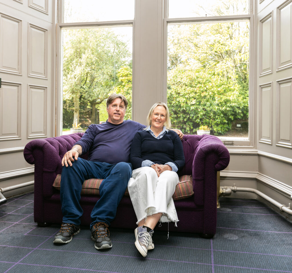 Campbell and Luci Carey sat in front of a large window and the Ardoe Hotel. Image supplied with release by Frame.