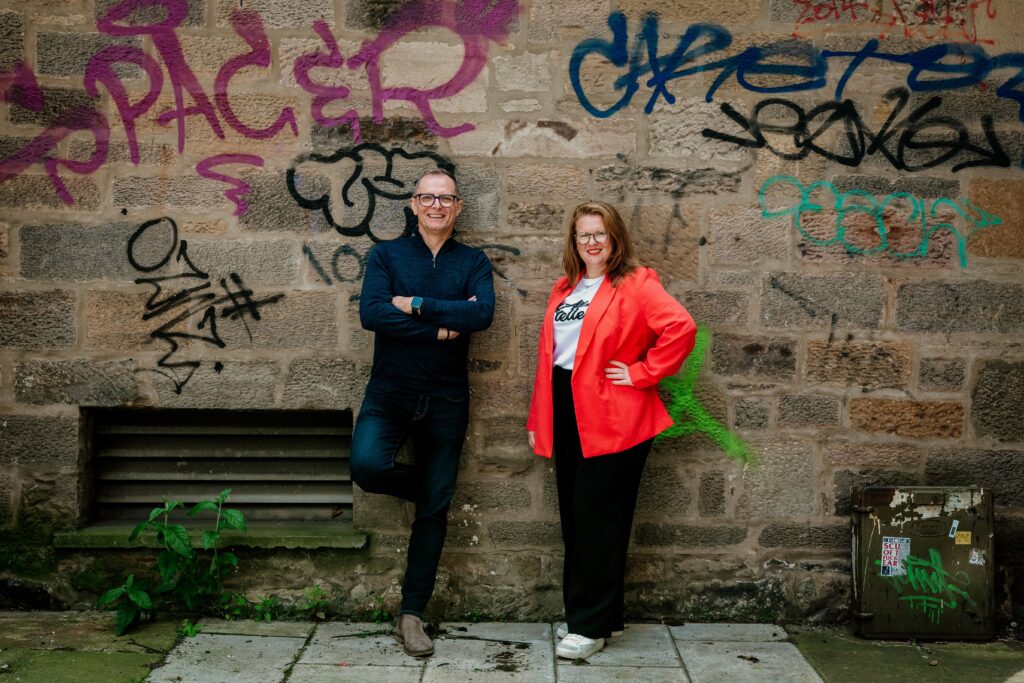 Kirsten Paul and Ray Bugg stand, smiling against a wall with graffiti. Image supplied with release by brass neck