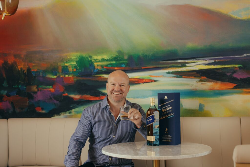 Scott Naismith enjoying a Blue Label in front of one of his landscapes in Bothy Bar. Image supplied with release by Frame Creates