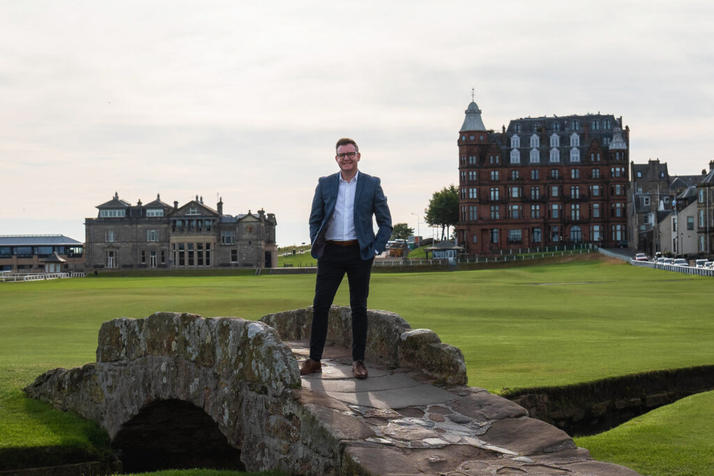 A man with dark hair and glasses, wearing a blue blazer, white shirt and black trousers, stands on top of a small stone bridge on a golf course.