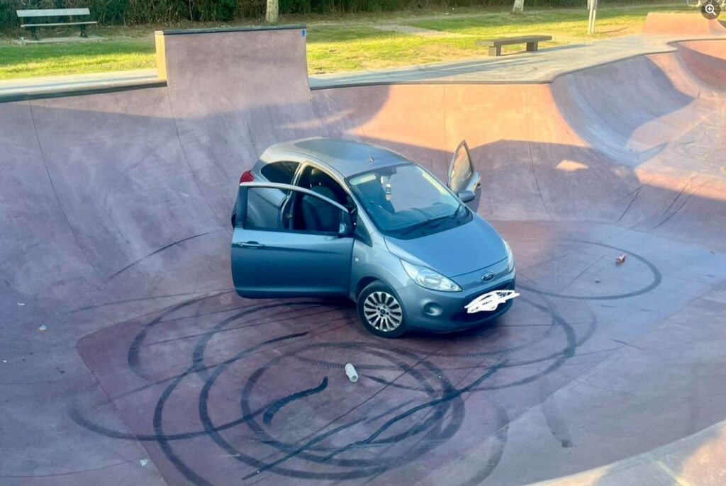 A grey Ford Ka abandoned at the bottom of a skatepark bowl with both front doors open. The ground surrounding it is covered in black skid marks.