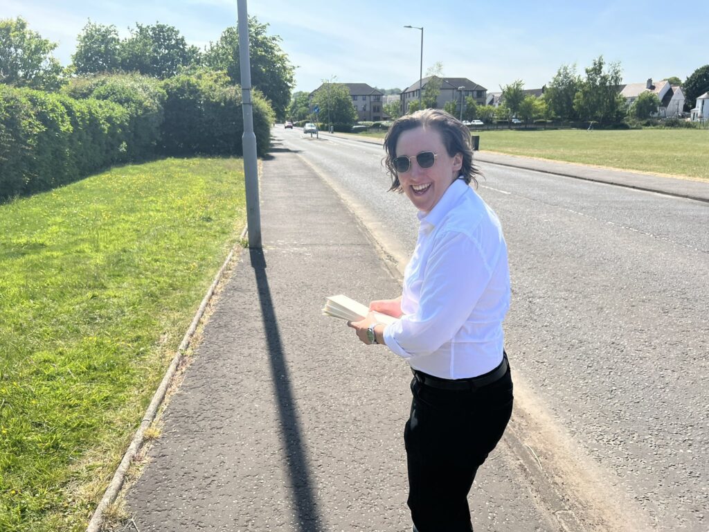 Scottish MP Mhairi Black wearing a white shirt and black trousers, standing in the street holding leaflets.