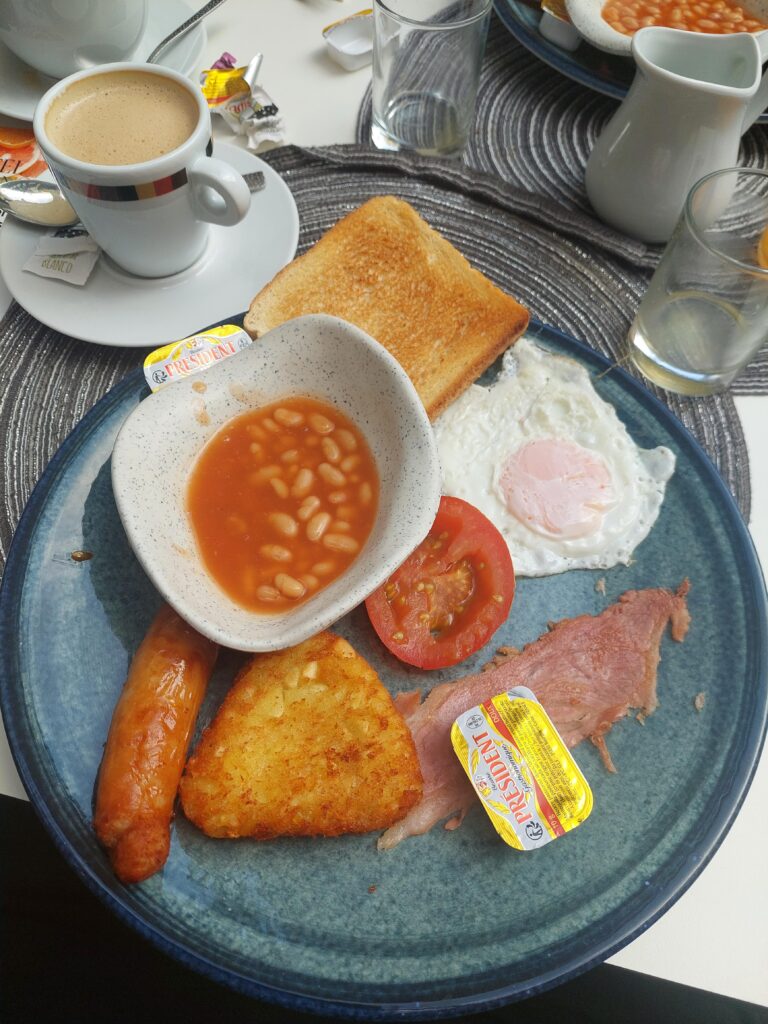 A blue plate with a messily-arranged full English breakfast: one fried egg, one tiny rasher of bacon, one sausage, one hash brown, one slice of toast and half a tomato. There is also a dish with a small serving of baked beans, two mini packets of butter sit at opposite sides of the plate.