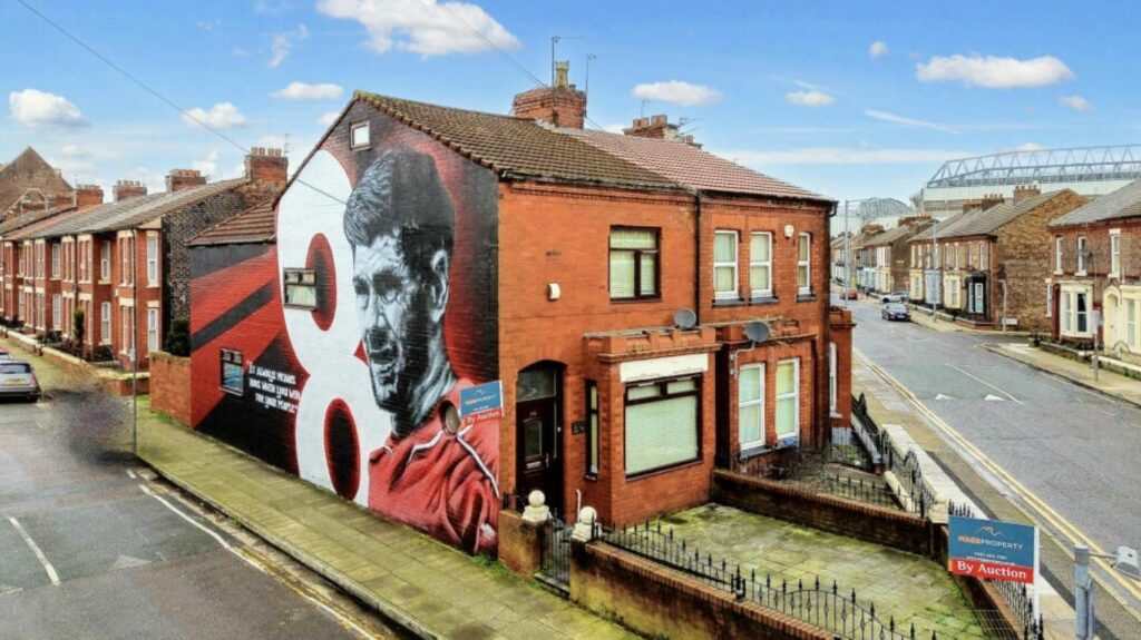 A picture showing the front and side of the house. The entire exterior side wall is painted red and black, with a likeness of Steven Gerrard running from the pavement to the roof,, a white number eight, and a quote in white lettering reading: "It always means more when you win for your people".