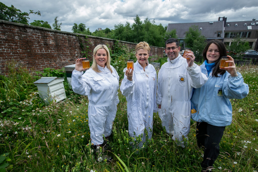 The hotel staff and hospice CEO, wearing beekeeper suits, and the beekeeper, wearing a light blue jacket, standing in long grass next to beehives and holding up jars of honey.