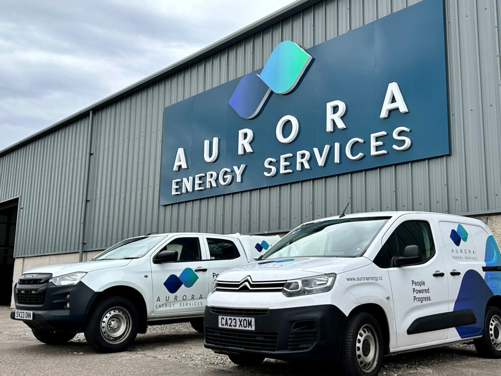 A pickup truck and a white van parked in front of a building with a sign reading "Aurora Energy Services" and a logo which resembles two abstract blue diamonds. Both vehicles feature the same branding.