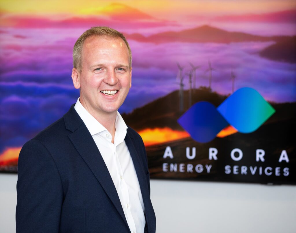 An image of a smiling man wearing a navy suit and white shirt. He is standing in front of a wall bearing a picture of a wind farm on a hill under a red and purple cloudy sky. The sign is branded with "Aurora Energu Services" and a logo which resembles two abstract blue diamonds.