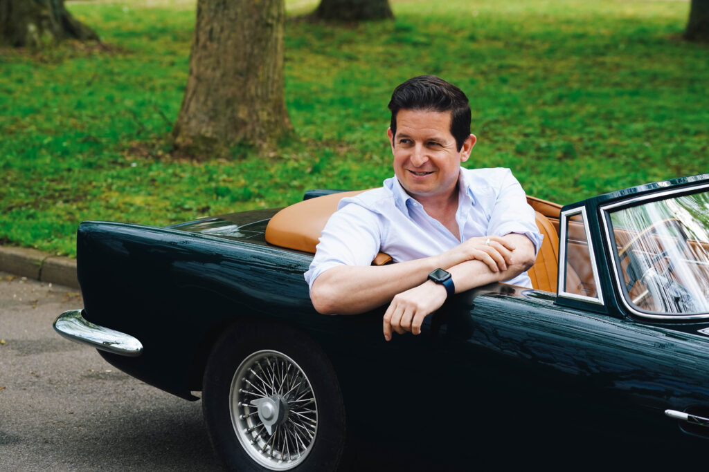 A dark-haired man wearing a white shirt sits in a scaled-down model of a black classic car.