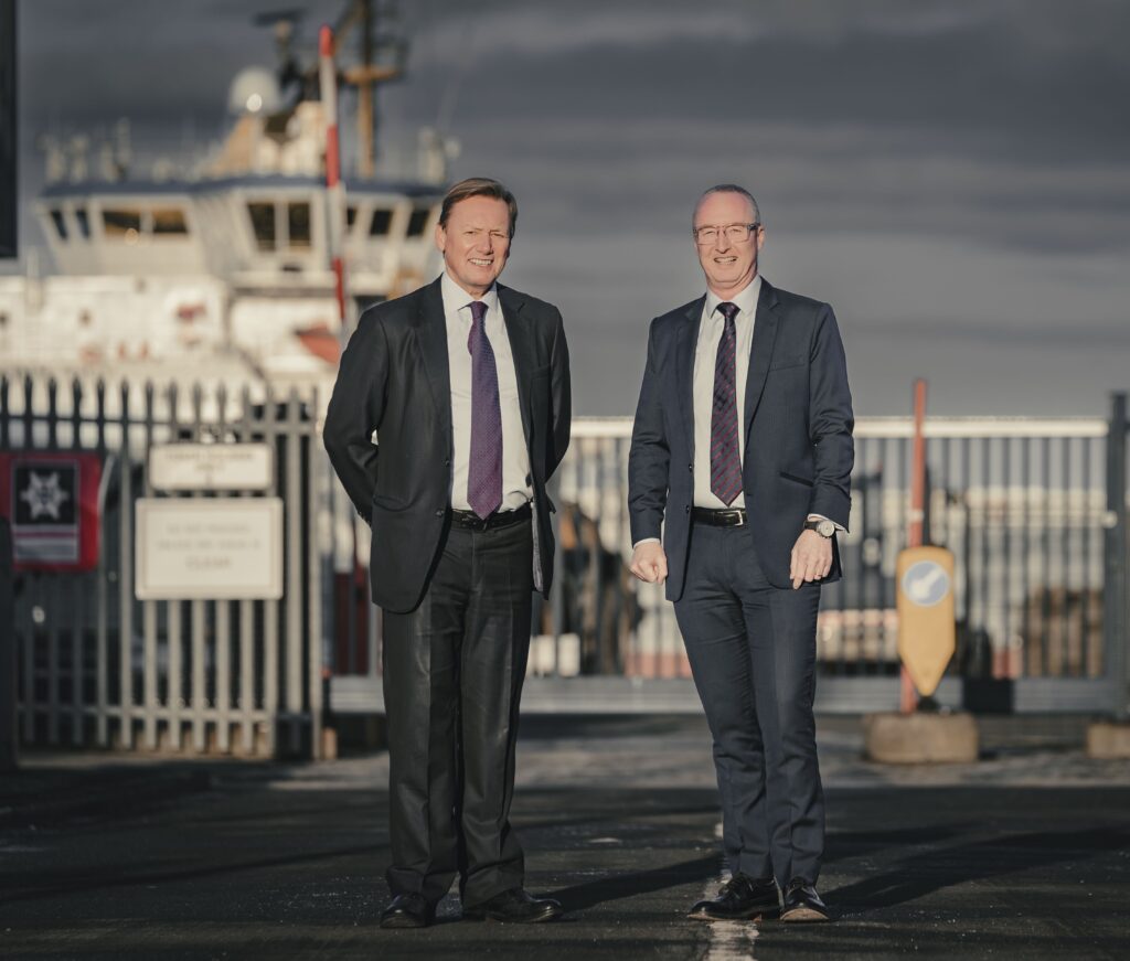 Two men, one dark-haired and wearing a black suit and purple tie, the other grey-haired with glasses and wearing a grey suit and black and red striped tie, stand in front of a metal fence, behind which is a marine vessel.