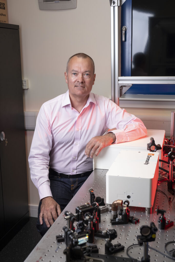 A grey-haired man wearing jeans and a pink shirt sits at a metal desk topped with lasers and technological equipment.