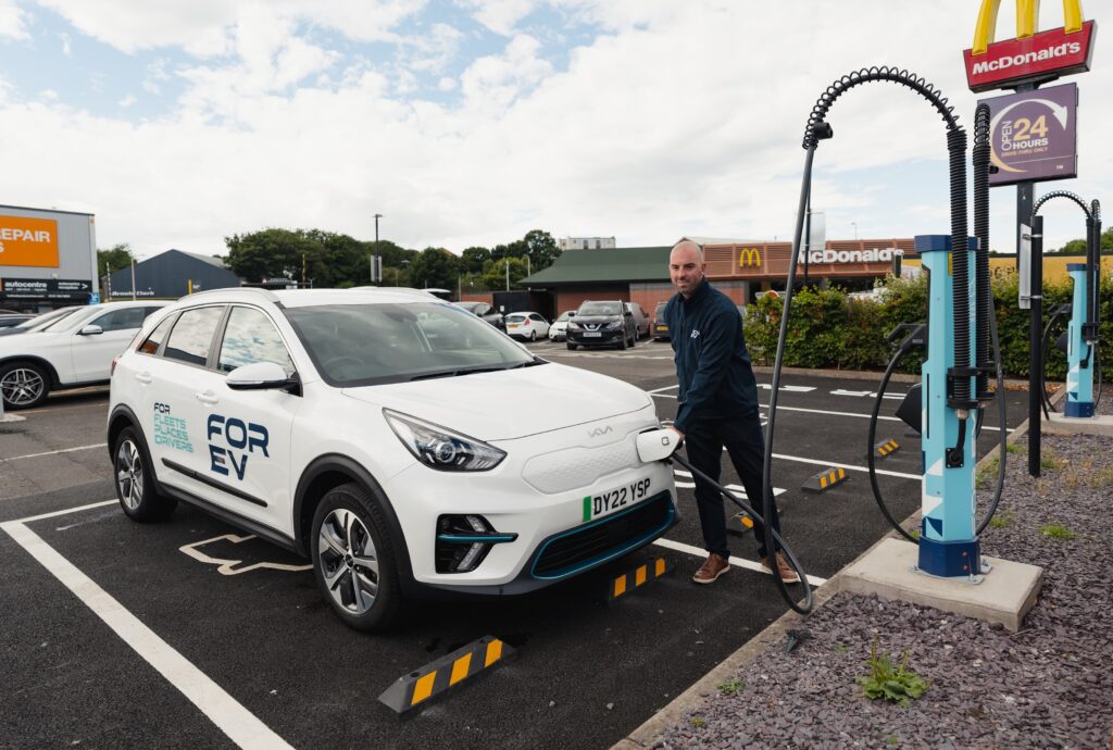 A man wearing a navy jacket and black trousers standing in a car park, plugging a charger into a white electric car branded with "FOR EV". 