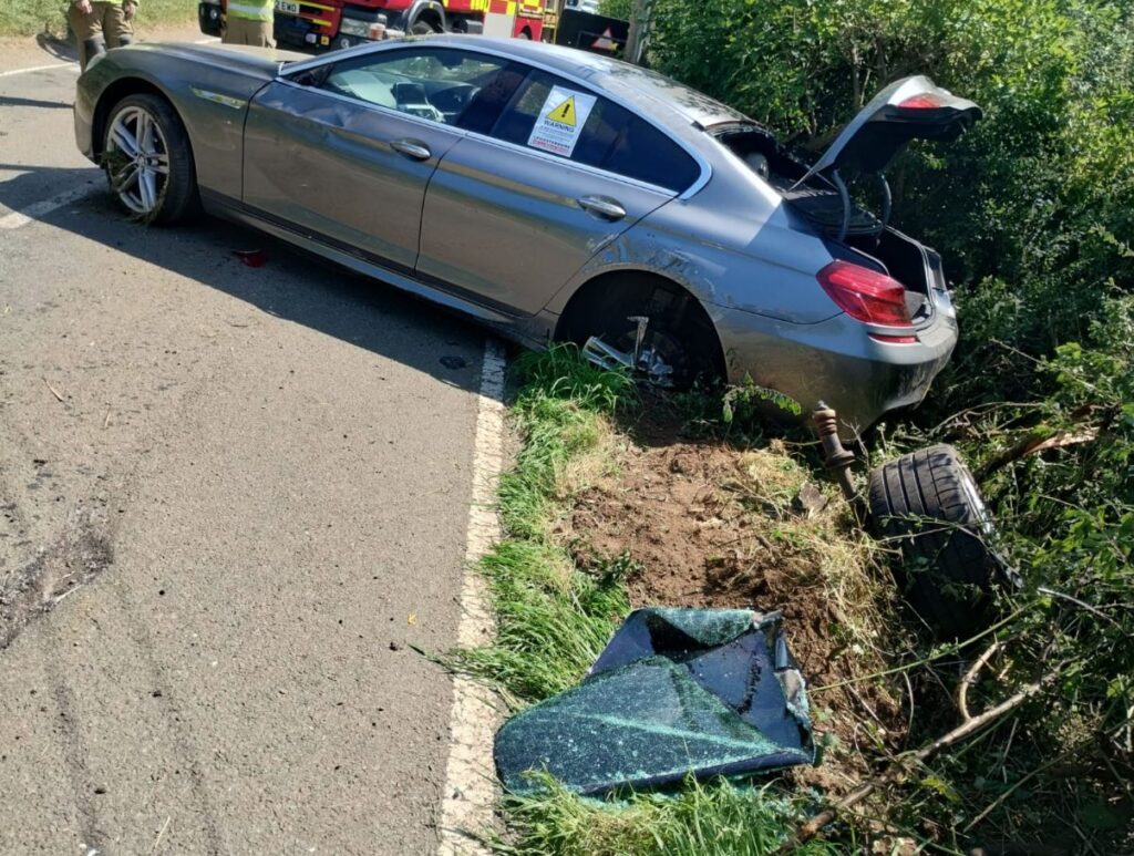 A silver BMW stopped at an angle on a country road, its back end in a hedge. The car is covered in dents and scratches, and the visible back wheel is buckled and almost completely ripped off, the tyre visibly discarded next to it.