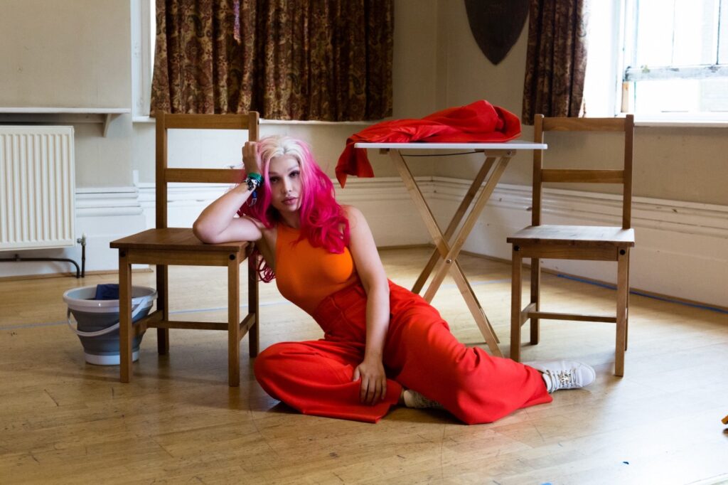 A woman wearing a blonde and pink dip-dyed wig, orange vest, red trousers and white trainers sits on a wooden floor leaning against a wooden chair. A table, holding a red jacket, and another chair are visible to her right, and there is a bucket on the floor to her left.