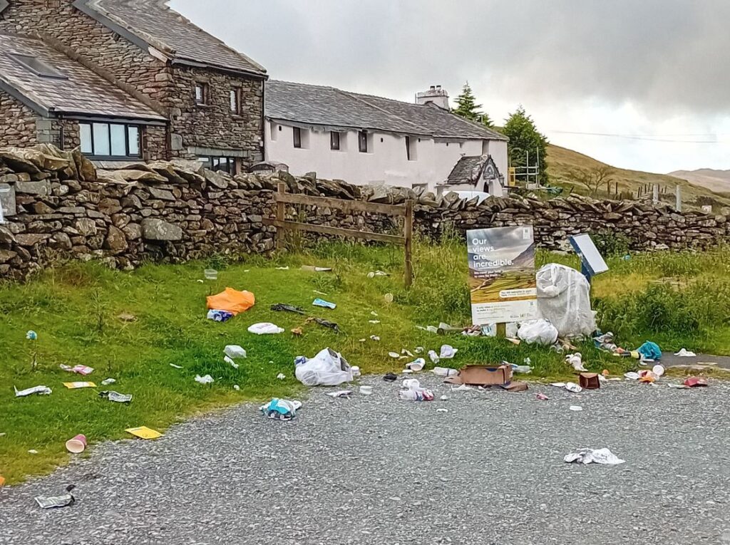 An image showing the edge of a footpath surrounded by grass and bordered by a stone wall, behind which cottages and hills are visible. A sign sits in the middle of the grass, reading: "Our views are incredible. We need your help to care for them." The grass and path are both covered with litter and bulging plastic bags strewn about.