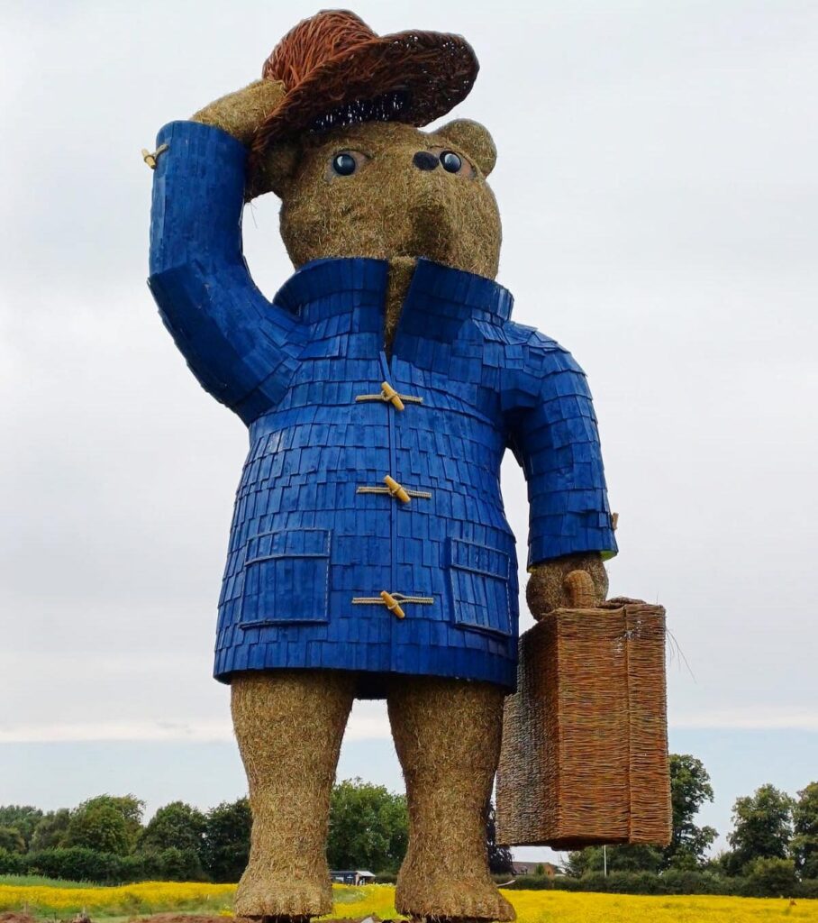 A giant straw and metal sculpture of Paddington Bear, a brown bear wearing a bright blue duffel coat. It raises a red hat with its right hand and holds a suitcase in its left hand. It stands in a field with trees and cloudy sky behind.