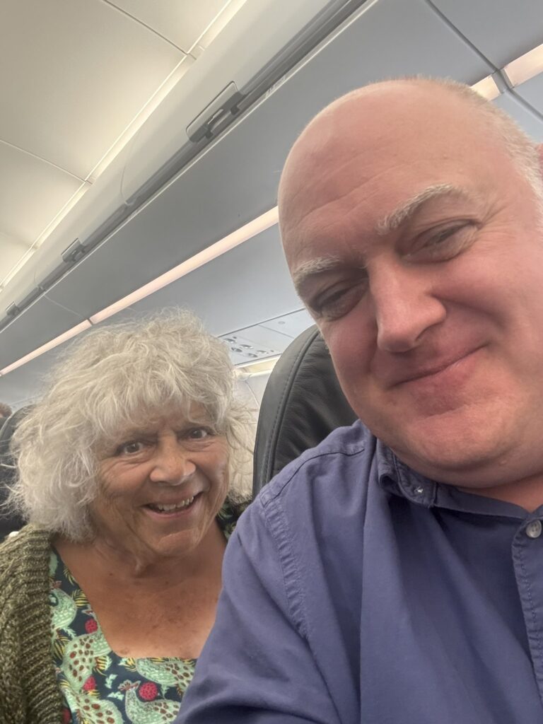 A selfie of two people sitting on board a plane. A man with thinning white hair, wearing a blue shirt, sits in front of a woman with curly grey hair, wearing a green cardigan over a blue, green and pink patterned top. Both are smiling.