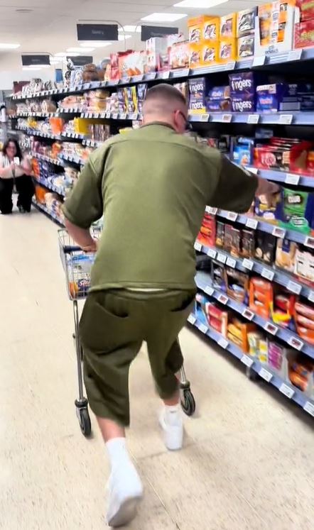 An image of a man with a shaved head and sunglasses, wearing a khaki green t-shirt and shorts, standing in a store pushing a shopping cart. He reaches for a shelf full of packets of biscuits to his right. A woman kneels at the end of the aisle and films him with her mobile phone.