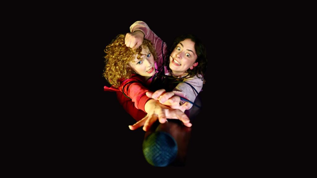 Two young women, one with curly dark hair wearing a light purple top, the other with curly red hair wearing a red jumper, reaching towards the camera for a microphone. The dark haired girl is pulling the red haired girl's hair.