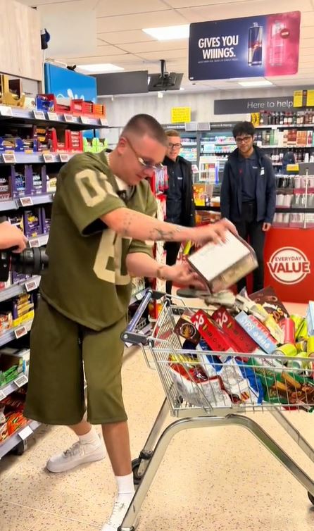 An image of a man with a shaved head and sunglasses, wearing a khaki green t-shirt and shorts, stands in a store. To his left are shelves of chocolate bars, and behind him are shelves of alcohol. He is tipping a box of chocolate bars into a shopping cart full of food and drinks.