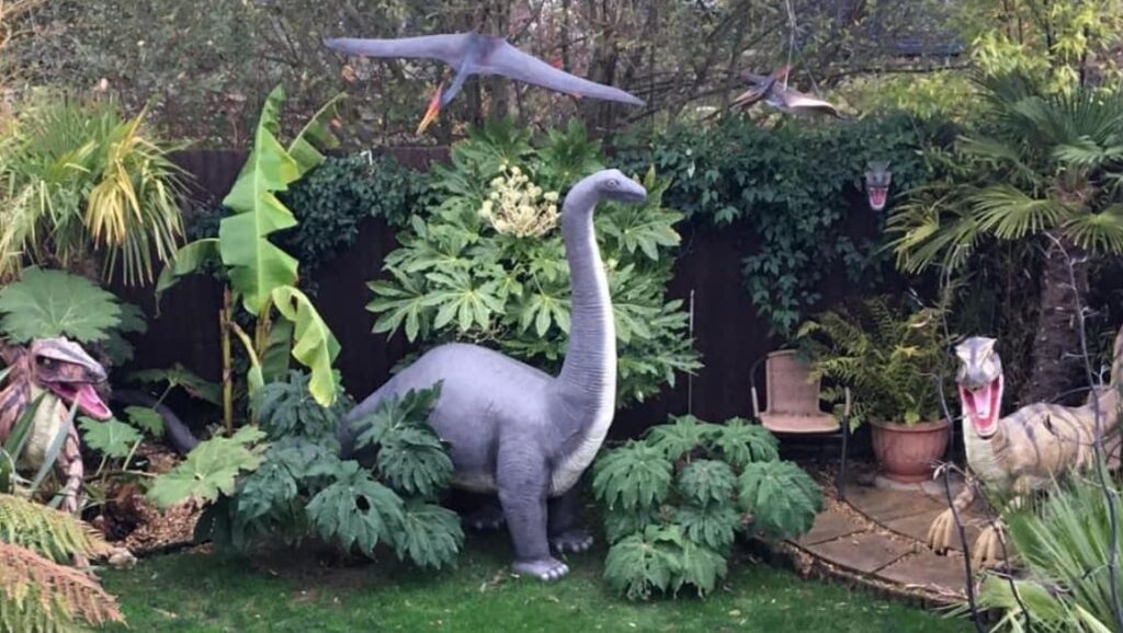 A garden with several green, leafy trees and shrubs, interspersed with large models of various dinosaurs, some on the ground, some hanging from trees.