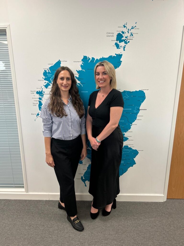 Two women stand in front of a blue map of Scotland on a white background. One has wavy dark hair, and wears a blue and white striped shirt, black trousers and black shoes. The other has short blonde hair and wears a long black dress and black heels.