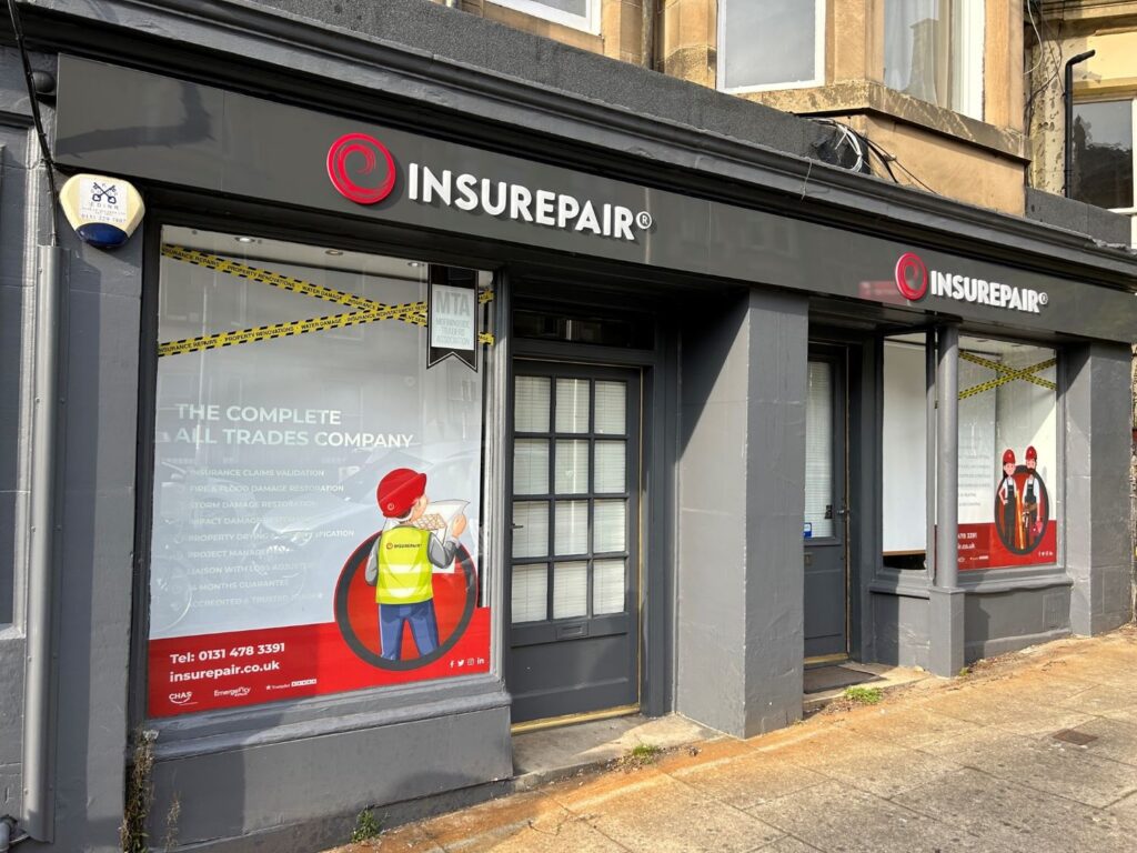 An image of the exterior of a building, with a grey shopfront, two windows and two doors. The name "Insurepair" is above both doors in white lettering next to a logo resembling a red spiral. Lettering in the window reads "The complete all trades company" and a list of services. A cartoon figure wearing a helmet and hi-vis jacket is also visible.