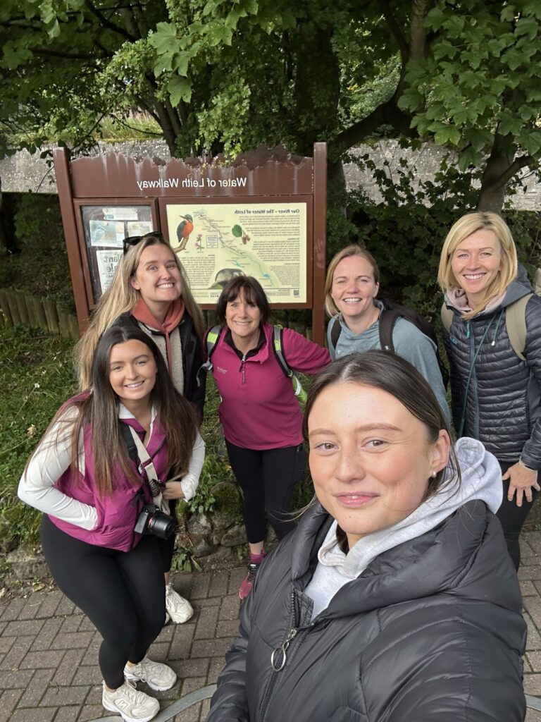 An image of a group of people dressed in outdoor clothing, standing on a path in front of a notice board.