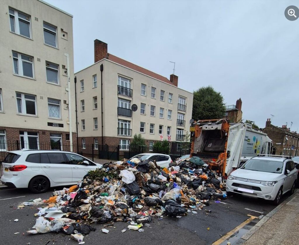 An image of a waste collection lorry, parked in the middle of the road, next to two blocks of flats. A huge pile of loose and bagged waste is piled on the road at the back of the lorry.