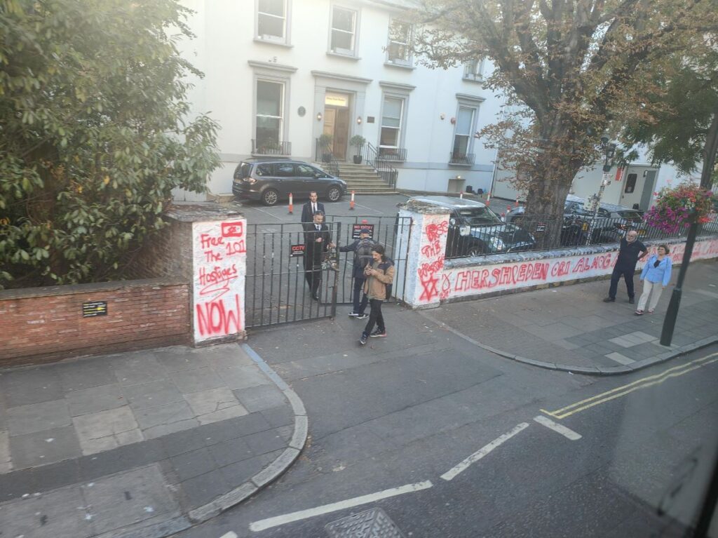 An image of a low white wall outside a building, almost completely covered in red spray painted letters and emblems.