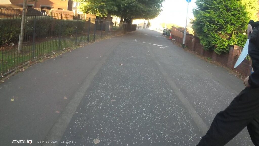 A still from a cyclist's helmet camera, showing a cycle path bordered by trees, black railings on one side and a low brick wall on the other side. A figure wearing black is just walking into shot, holding a realistic looking plastic knife,