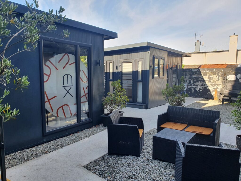 An image of seating on a patio outside two modern black shed-like buildings with tall glass windows.