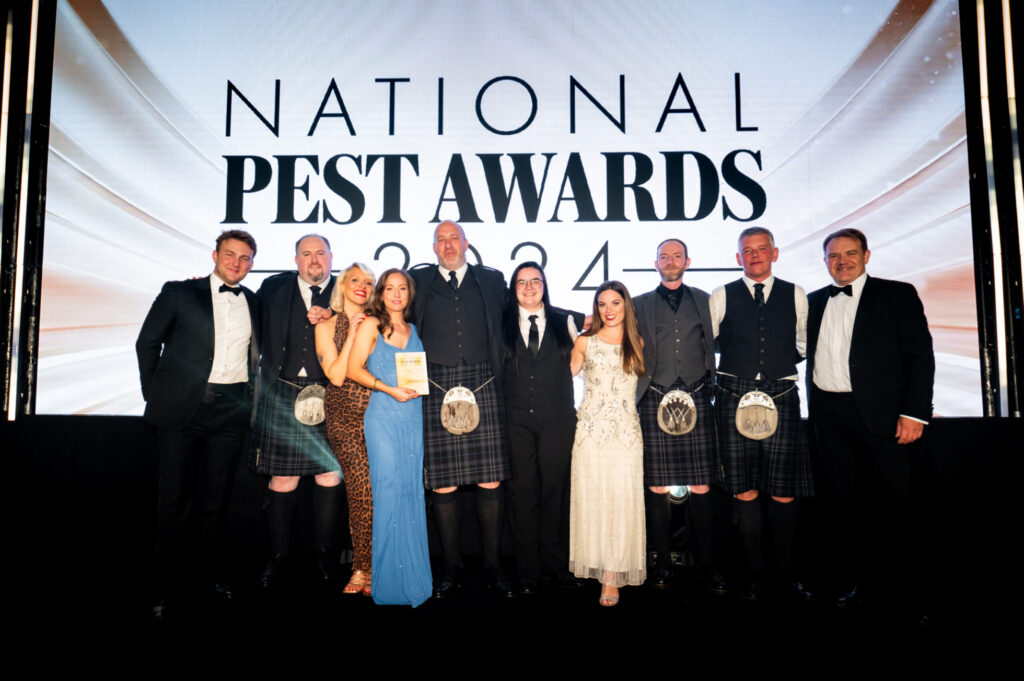 A group of ten people dressed in formal attire, standing in front of a screen which reads: "National Pest Awards 2024." A woman in a blue dress holds an award plaque.