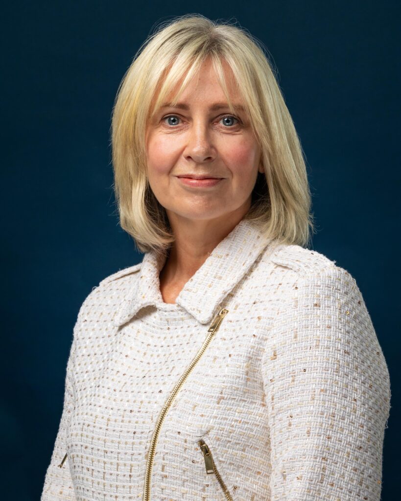 An image of a woman with shoulder-length blonde hair, wearing a white and gold jacket.