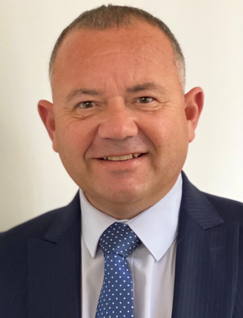 An image of a man with short grey hair, wearing a navy suit and blue and white spotted tie.