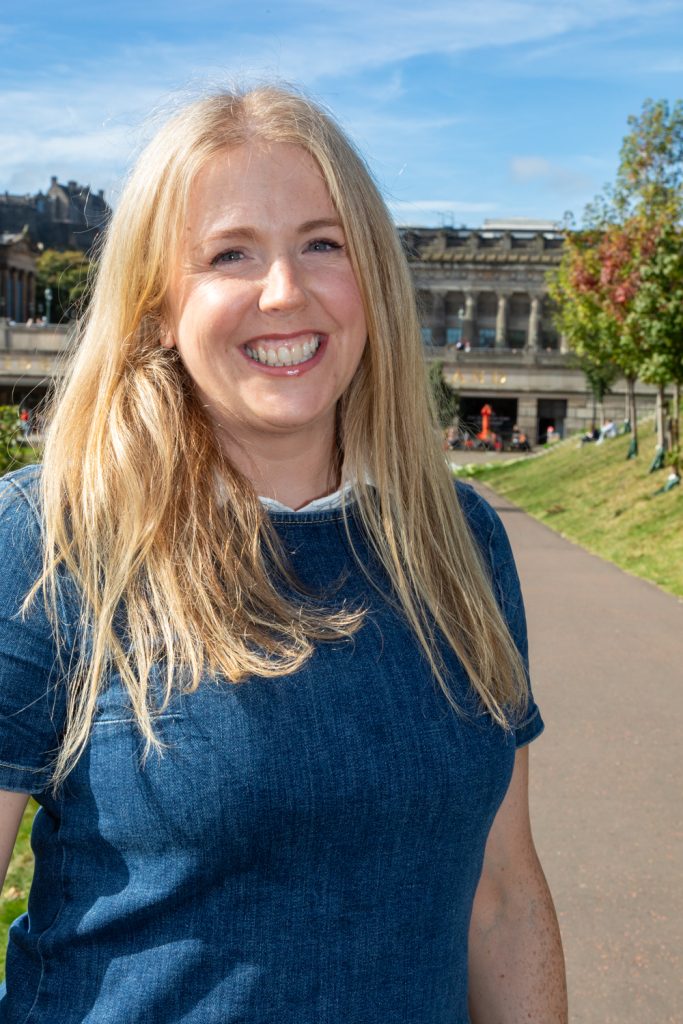 An image of a smiling woman with long blonde hair, wearing a blue top, standing on a footpath.