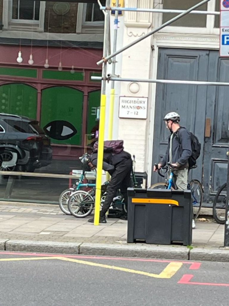 An image of two men wearing helmets, standing aside bikes chained up on a pavement. One appears to be tampering with the lock on one of the bikes.