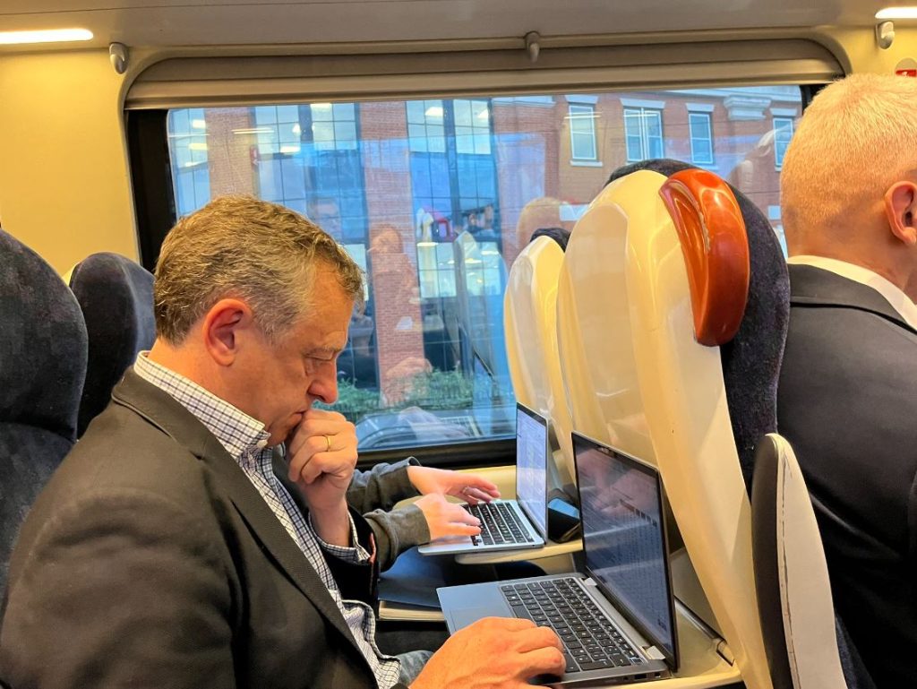 An image of a man in a suit sitting on a train, using a laptop. A second person is just visible next to him, also using a laptop.