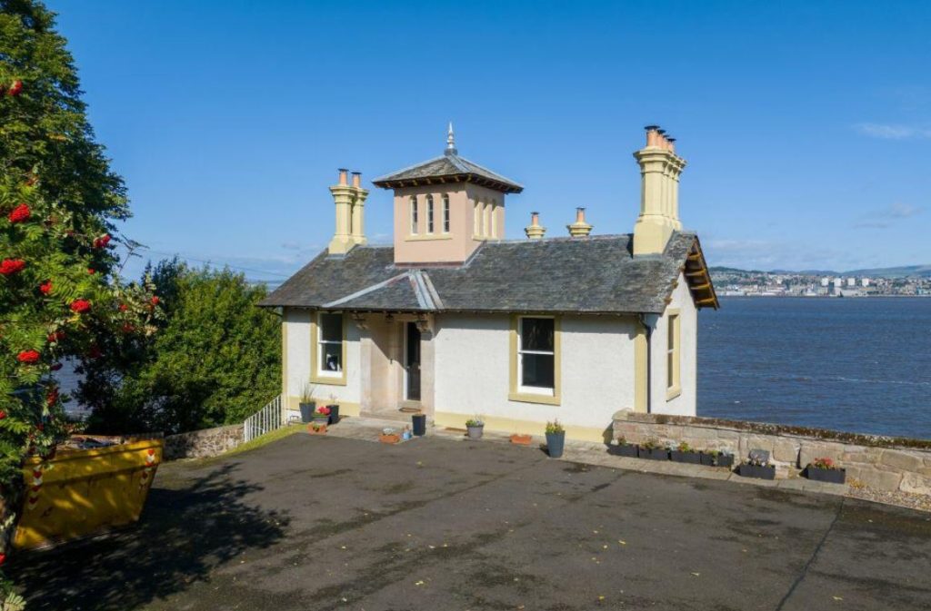 An image of a white house with a grey roof and yellow windows. It sits at the end of a driveway with a river behind and a city in the distance.