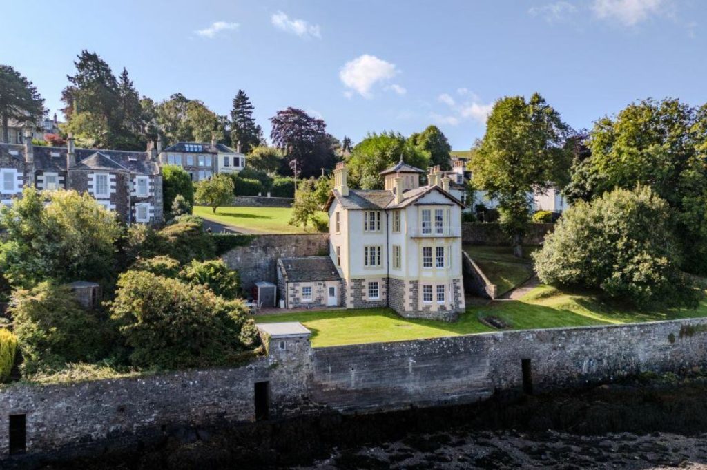 An image of a three-storey white house with a grey roof and yellow windows, positioned next to a river, with the bottom two storeys below street level. It is surrounded by trees and a few other houses.