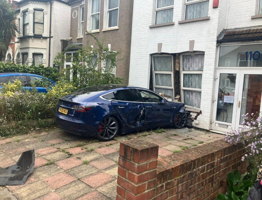 An image of a dark blue car crashed head-on into the front window of a white brick house.