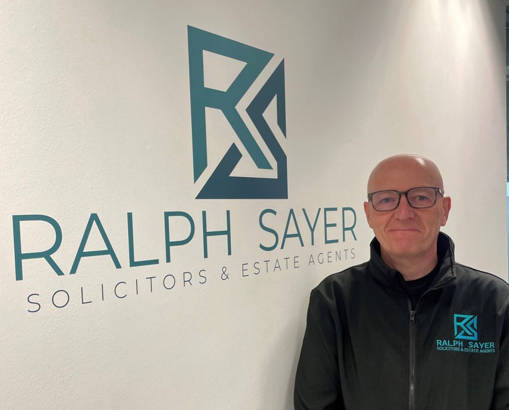 An image of a man wearing glasses and a black jacket with a blue logo. He stands next to a wall featuring the same logo and the name: "Ralph Sayer Solicitors and Estate Agents."