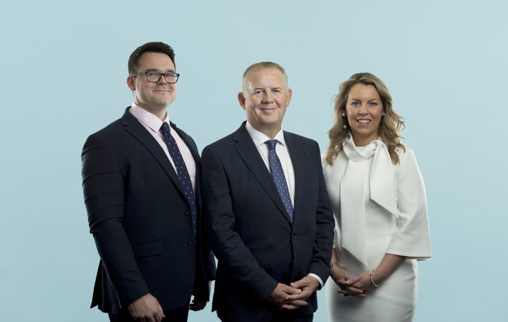 An image of two men and a woman in business attire, standing in front of a light blue background.