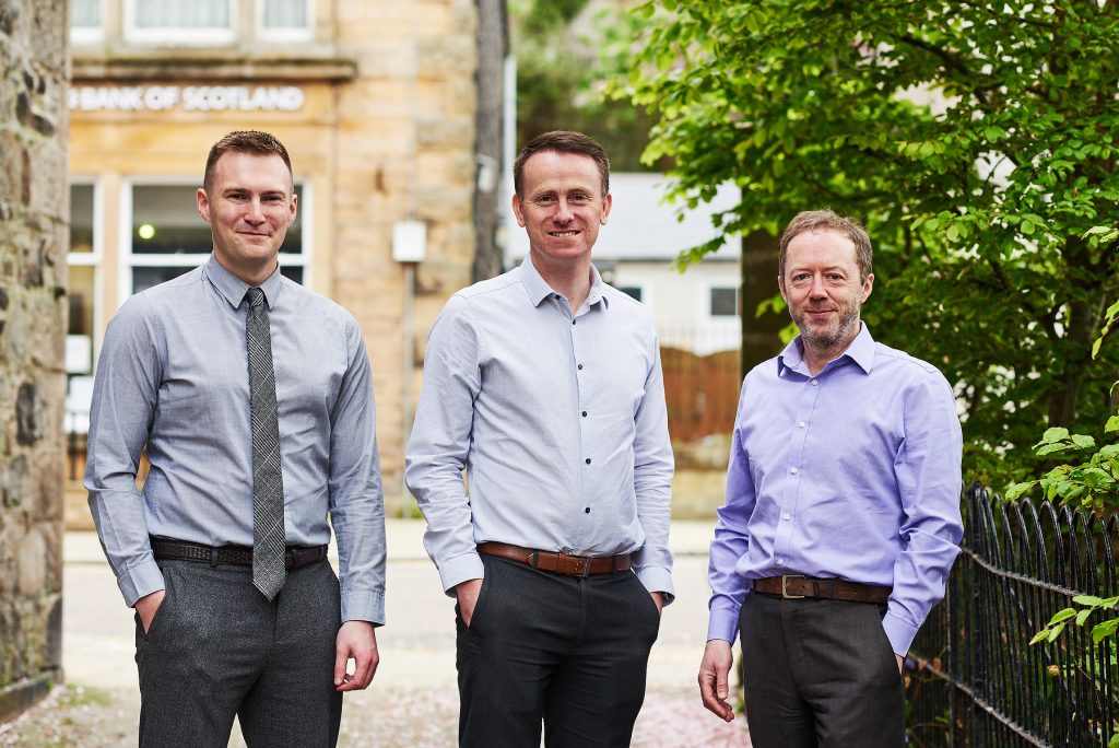 An image of three men in business attire, standing outside a stone building.