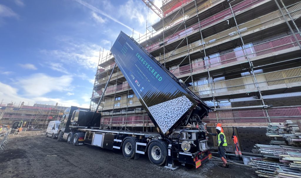 An image of a truck on a building site, tipping material into a machine.