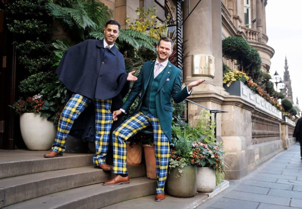 Two men outside a building wearing smart jackets with blue and yellow tartan trousers