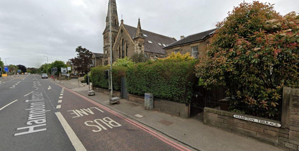 An image of a street, with houses and a church visible behind a high hedge.