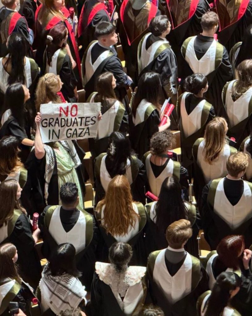 An image of a crowd of people wearing graduation robes, with one holding up  a sign reading: "No graduates in Gaza"