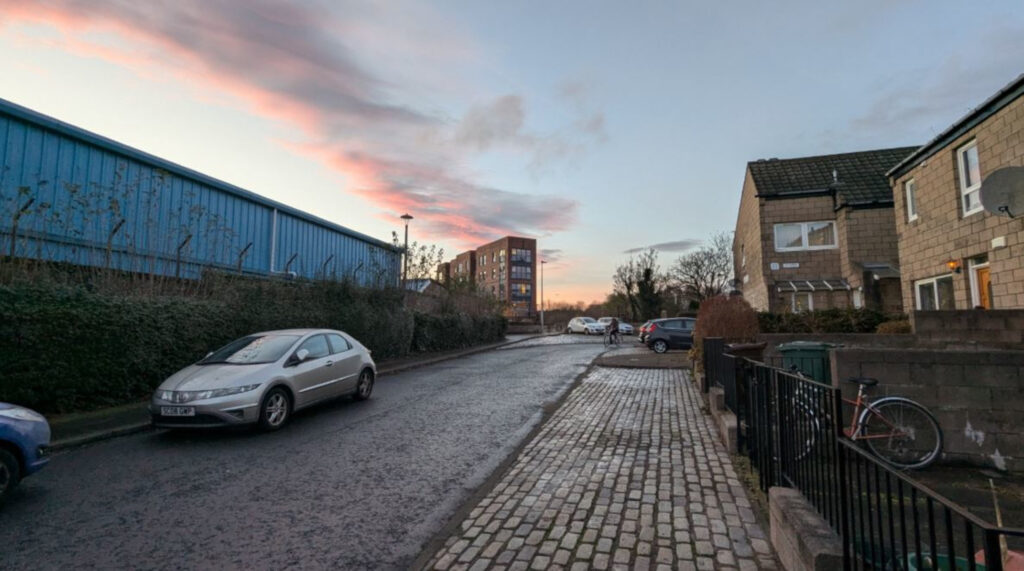 An image of a cobbled street, with a blue industrial building on the left and houses on the right.