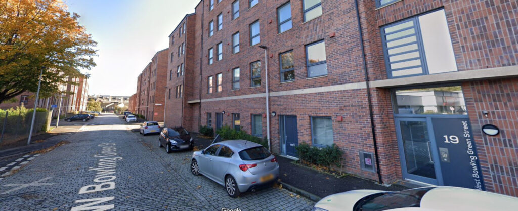 A image of a street with a red-brick building and parked cars down the right-hand side.