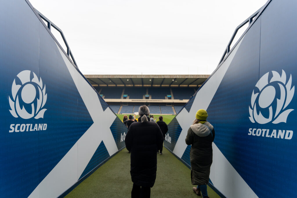 An image of people walking between two high-sided blue walls on to a rugby pitch.